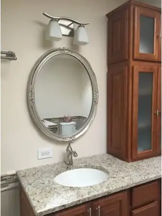 Oval mirror above a cherry bathroom cabinet with a stone top