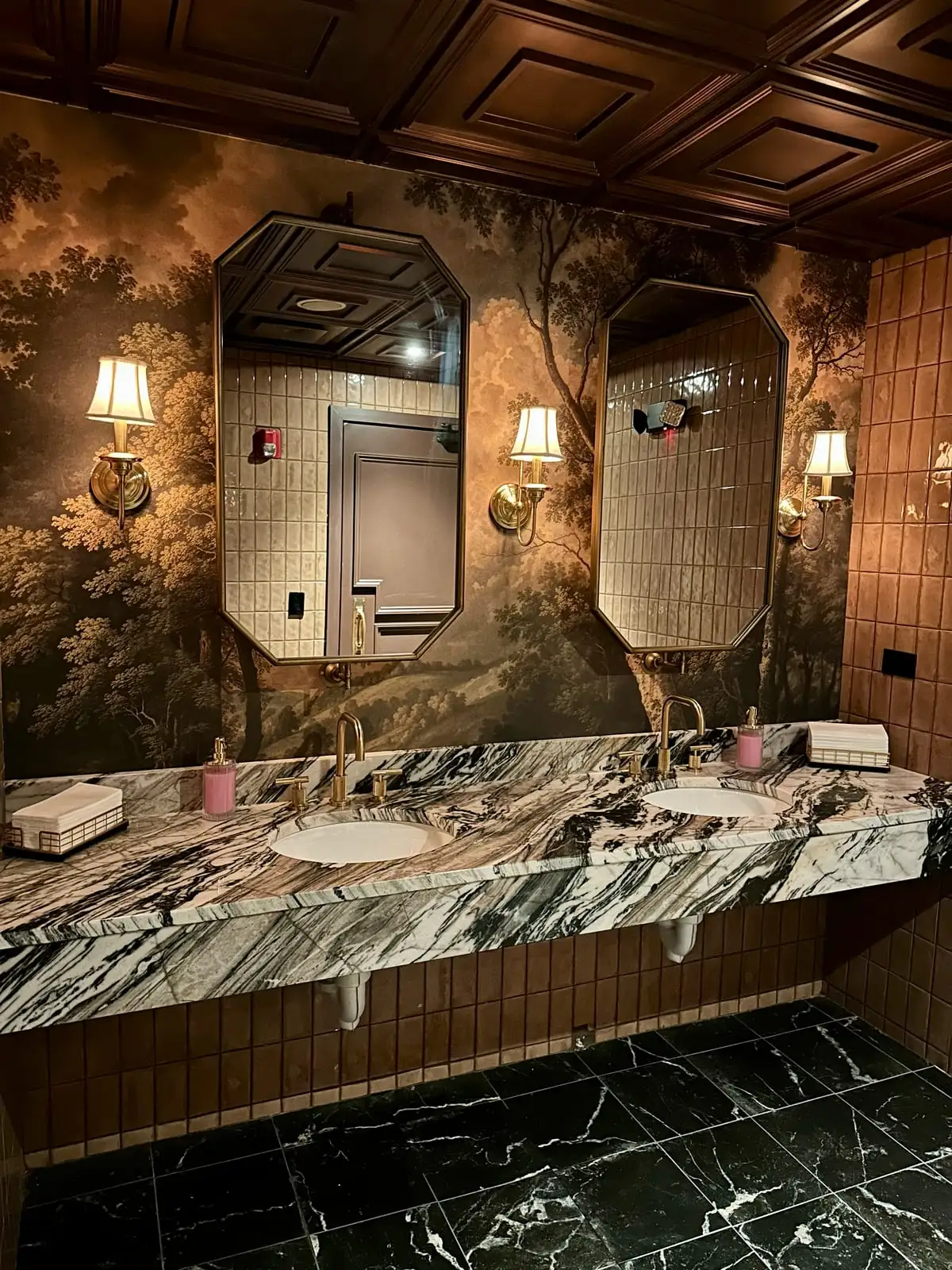 Restaurant restroom with a boldly veined marble vanity, forest-mural walls and brass sconces