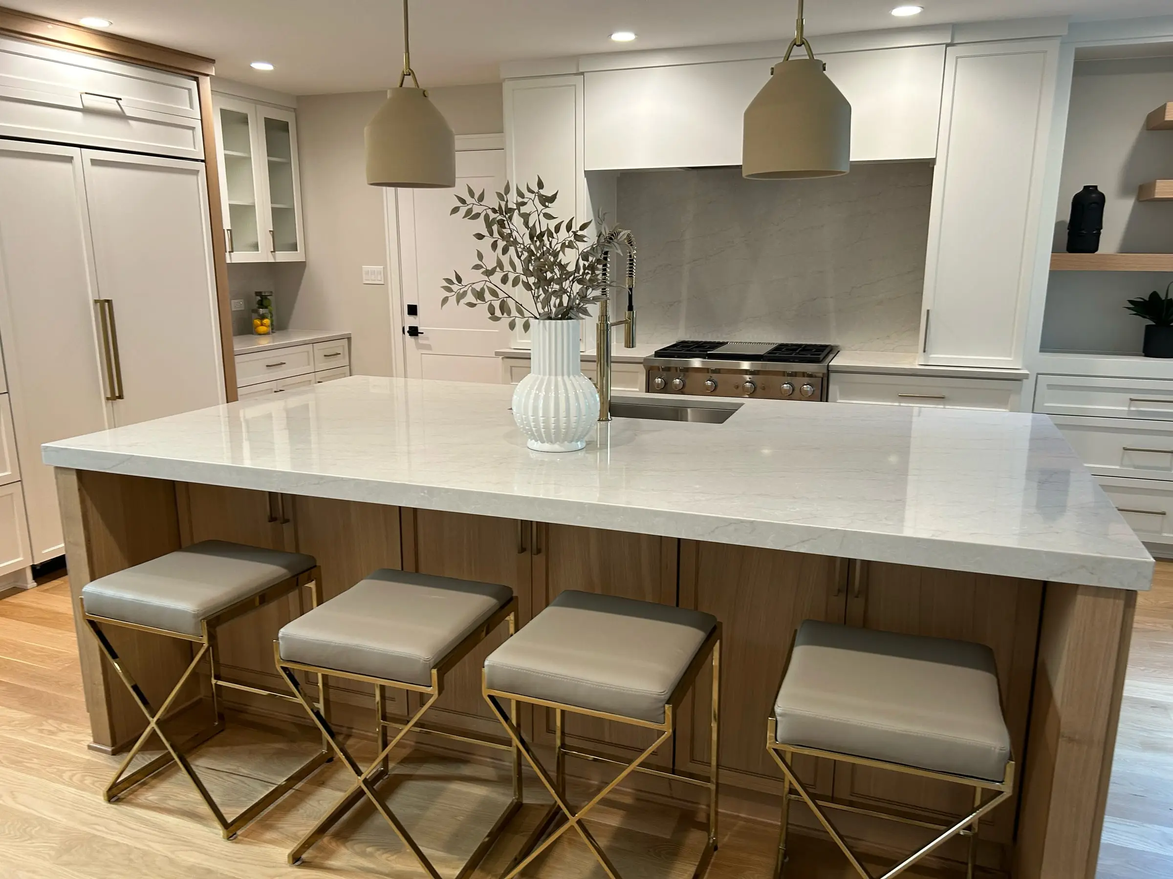 Bright white kitchen with an oversized quartz island, white-oak base and brass counter stools, fabricated and installed by The Granite & Marble Depot