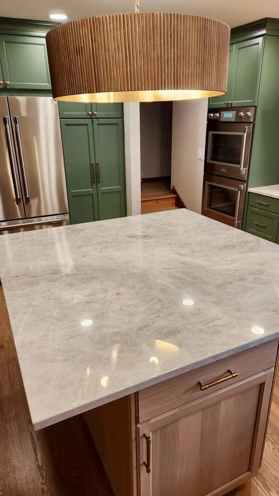 Deep green cabinets with a Taj Mahal quartzite island beneath a fluted brass drum light